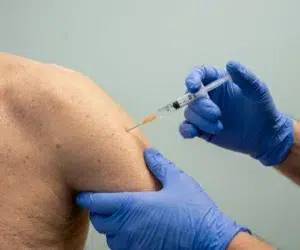 Braço de uma pessoa em close-up recebendo uma injeção de vacina. Apenas o braço e a seringa estão visíveis, enquanto um profissional de saúde com luvas azuis aplica a injeção com precisão. Iluminação neutra, foco nítido e textura de pele detalhada em ambiente clínico estéril, com fundo discreto.