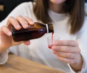 Pessoa despejando xarope com clobutinol medicinal em um copinho dosador, demonstrando a medida correta para consumo.