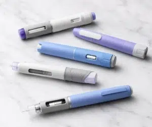 Conjunto de cinco canetas injetáveis para emagrecimento dispostas sobre uma superfície clara com acabamento marmorizado. Os dispositivos possuem design moderno nas cores branca, azul e lilás, com detalhes metálicos e visores transparentes, fotografados em alta resolução sob iluminação suave.