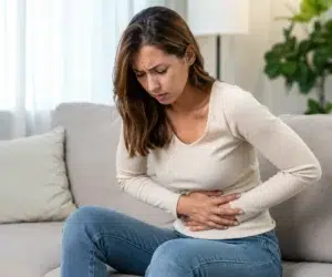 Mulher adulta sentada em um sofá claro, inclinada para frente e segurando a região inferior do abdômen com as duas mãos, demonstrando expressão de dor abdominal. O ambiente é interno, bem iluminado e desfocado ao fundo, com almofadas, cortinas, uma luminária e uma planta decorativa. A imagem é realista e ilustra desconforto ou cólica abdominal.
